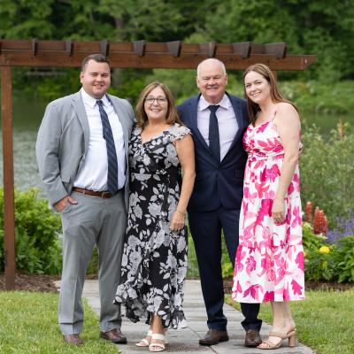 Four people pose together by a lake, smiling in formal attire amidst greenery.