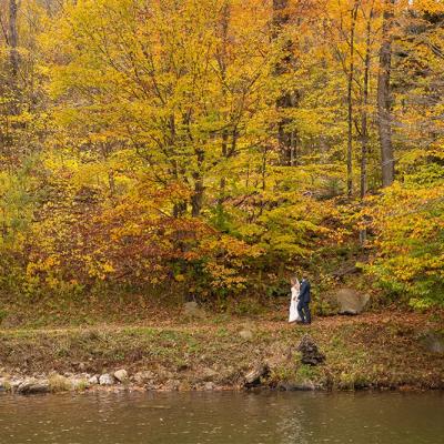 A couple stands by a river surrounded by vibrant autumn foliage.