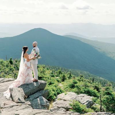 A couple in wedding attire stands on a rock, surrounded by mountains and greenery.