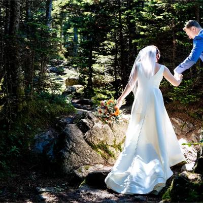A bride in a white gown and groom in a blue suit walk hand-in-hand through a forest trail.