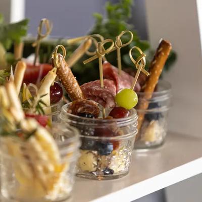 Mini jars filled with snacks, including fruits, meats, and pretzels, garnished with herbs.