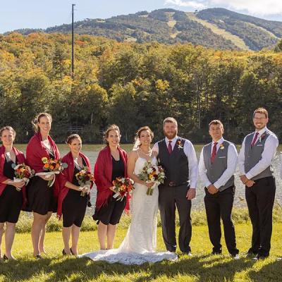 Bridal party posing by a lake with autumn foliage and mountains in the background.