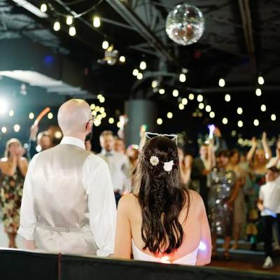 A couple stands together at their wedding reception, surrounded by cheering guests.