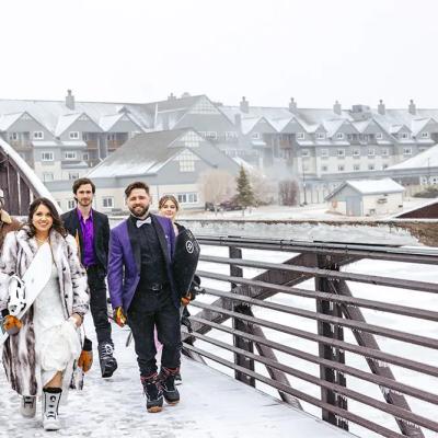 Group of friends in winter attire walking on a snowy bridge, snowboarding gear in hand.