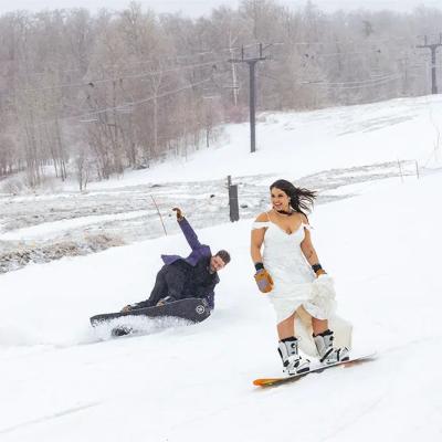 Bride snowboarding down a hill while a friend tumbles on a snowboard in the snow.
