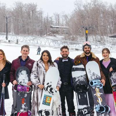 Group of eight people in winter attire, holding snowboards at a snowy ski resort.