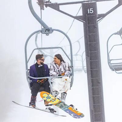 A couple rides a ski lift in snowy weather, dressed warmly, and smiling at each other.