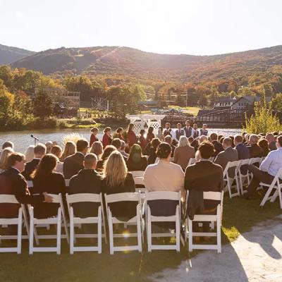 Outdoor wedding ceremony with guests seated by a lake, surrounded by autumn foliage.