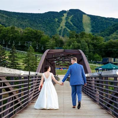 A bride and groom walk hand in hand across a scenic bridge surrounded by mountains.