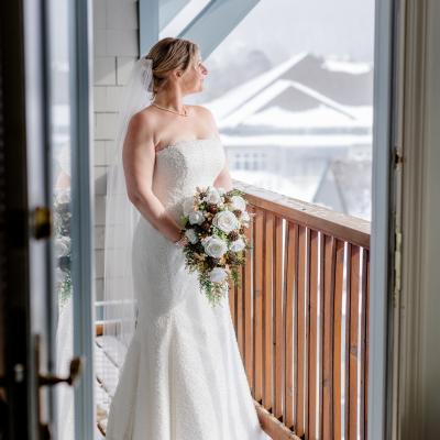 Bride in a strapless white gown stands on a snowy balcony, holding a floral bouquet.