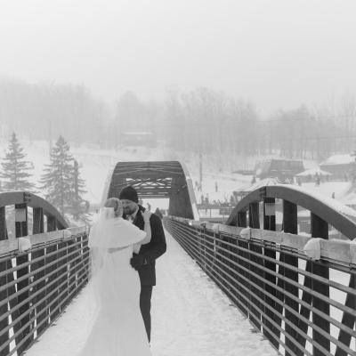 A couple in wedding attire embraces on a snowy bridge in a foggy landscape.