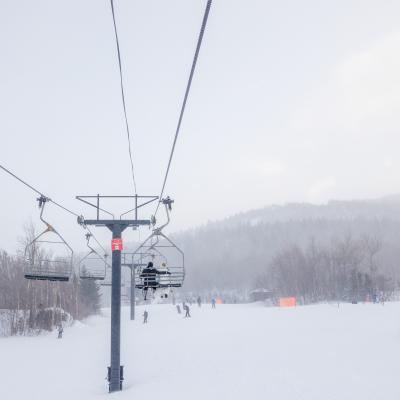 Ski lift over a snowy slope with skiers in the distance and foggy mountains.