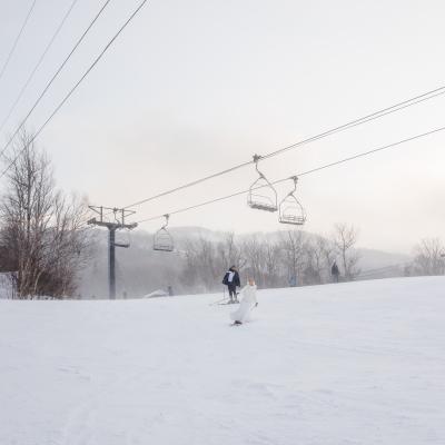 A skier in white attire glides down a snowy slope with a ski lift in the background.