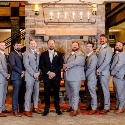 Groomsmen in grey suits stand in a lineup by a stone fireplace, smiling.