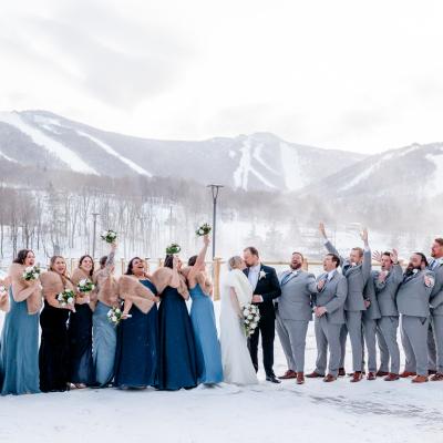 A joyful wedding party in winter attire poses in the snow with mountains in the background.