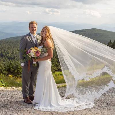A happy couple in wedding attire poses outdoors with mountains in the background.