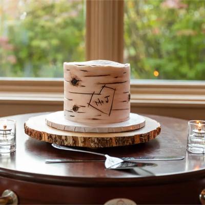 A rustic wedding cake on a wooden stand, engraved with initials M and J, beside candles.