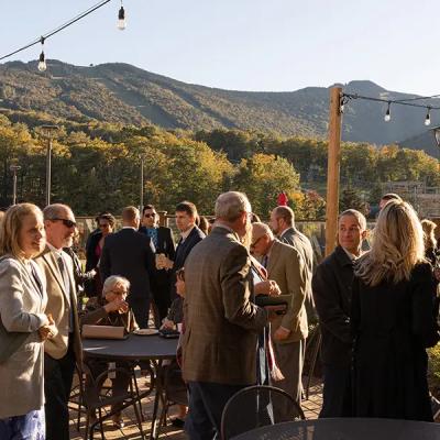 A crowd mingles outdoors at a scenic venue with mountains in the background.