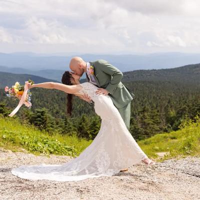 A bride in a lace gown and groom share a romantic kiss against a scenic mountain backdrop.