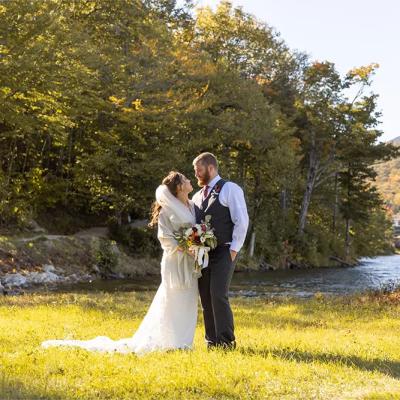 A couple in wedding attire embraces by a serene lakeside, surrounded by colorful autumn trees.