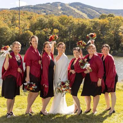 Bridal party by a lake, wearing black dresses and red shawls, holding bouquets.