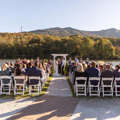 Outdoor wedding ceremony by a lake, surrounded by mountains and an audience in chairs.