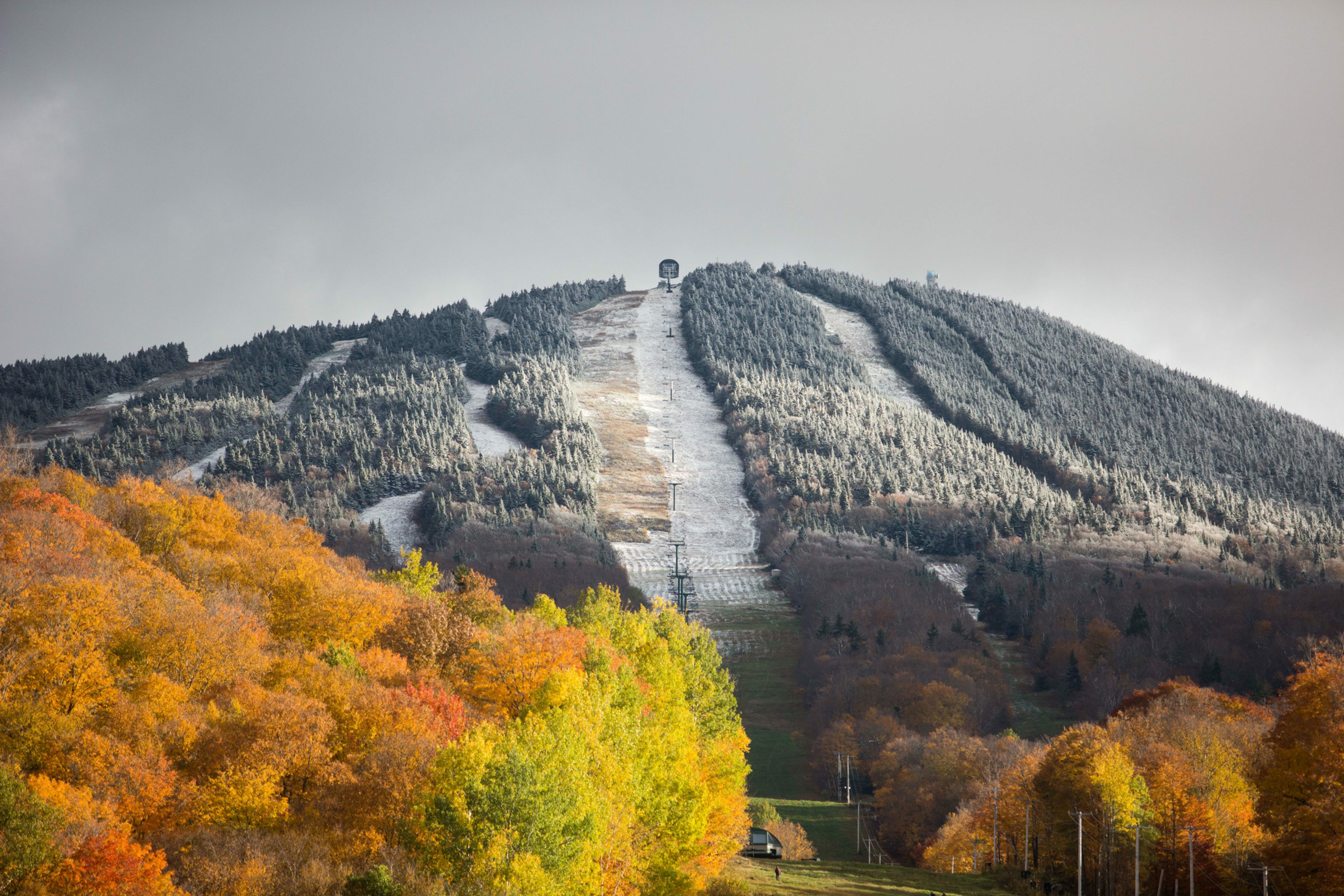 Mountain covered in autumn foliage and a snowy ski slope under a cloudy sky.