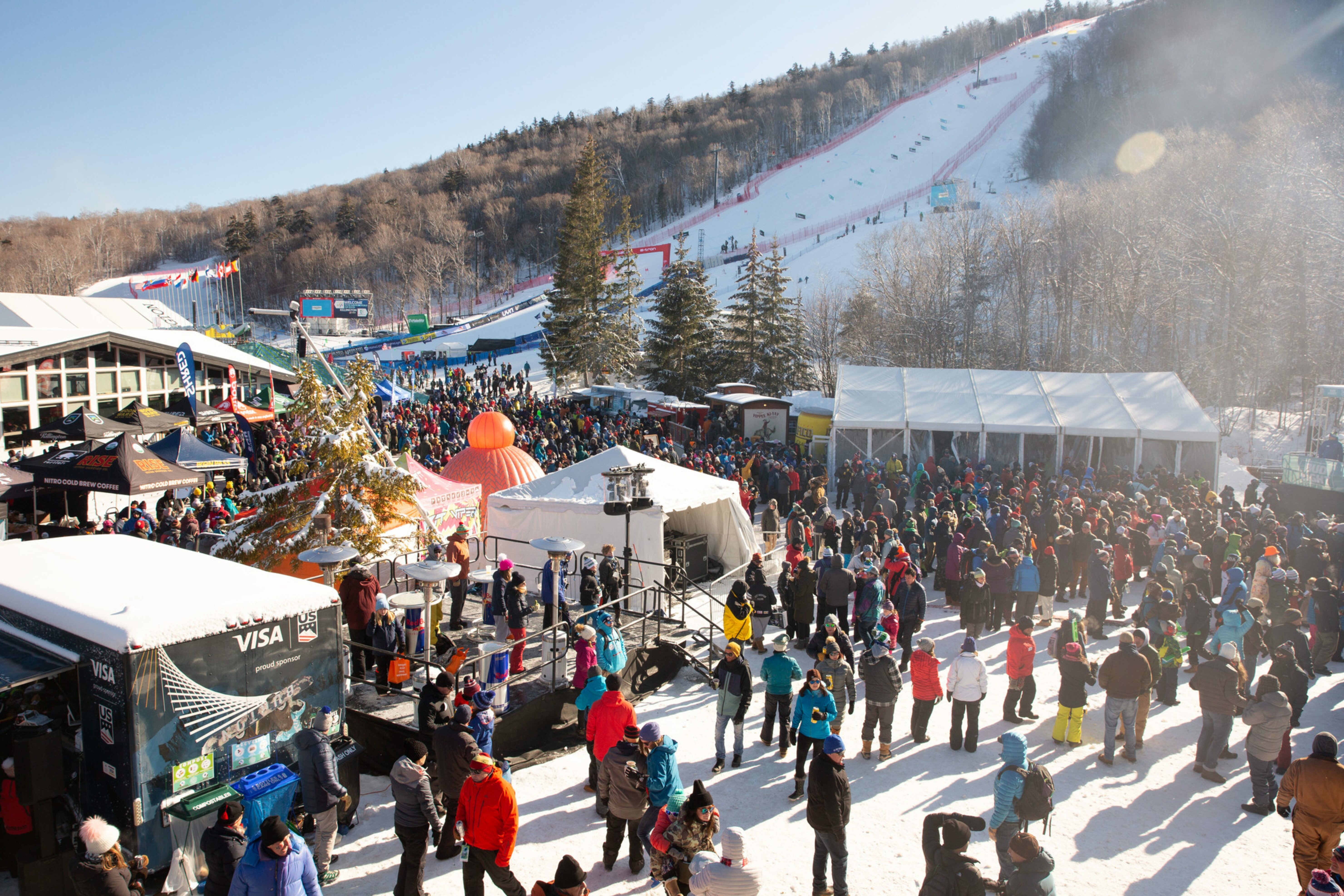 Crowd enjoying a winter festival at a ski slope, with food tents and a stage.