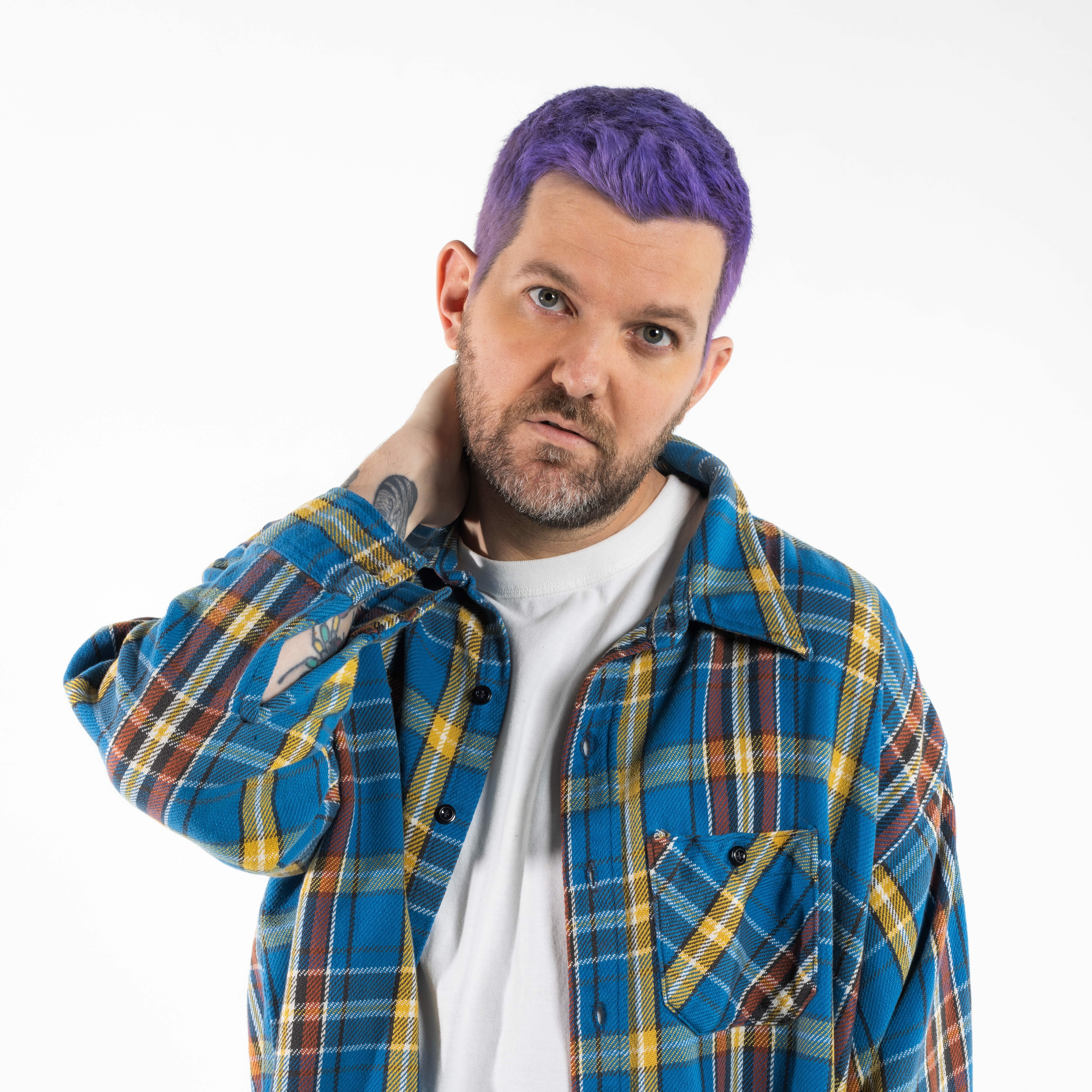 Person with purple hair wearing a plaid shirt poses thoughtfully against a white background.