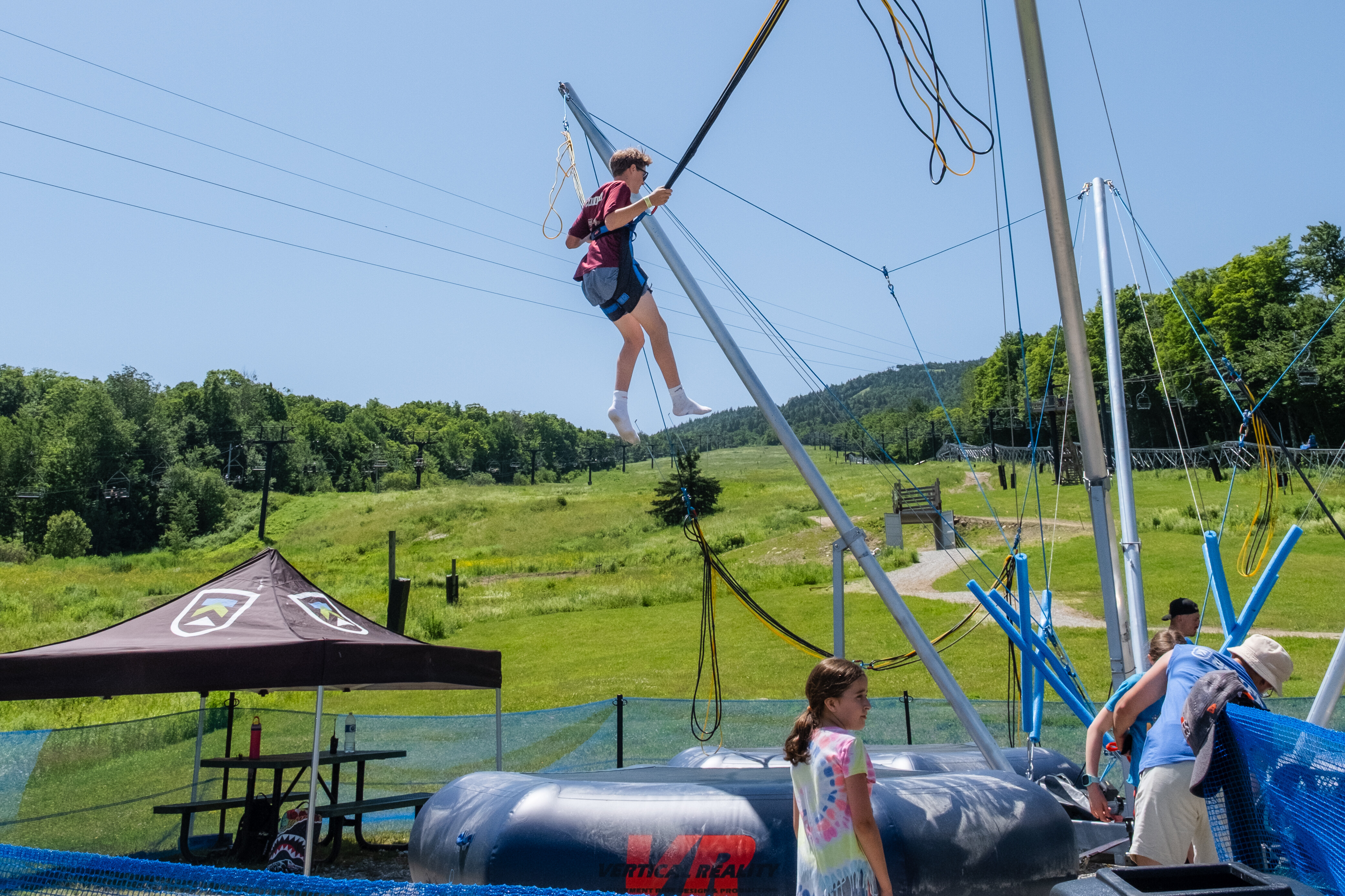 Child bouncing on a trampoline harness, with mountains and a summer sky in the background.