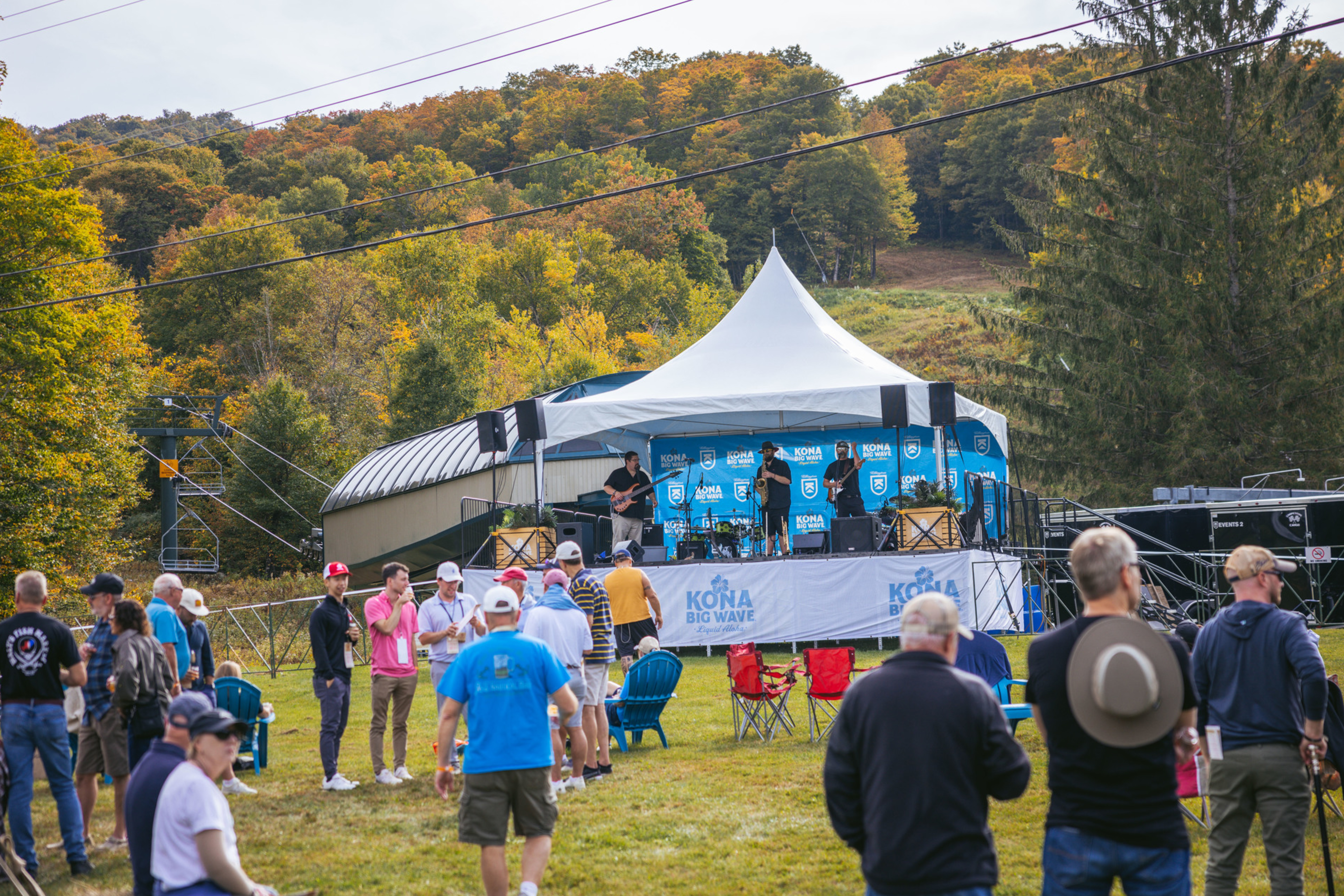 A crowd gathers near a stage with musicians performing outdoors in a scenic autumn setting.