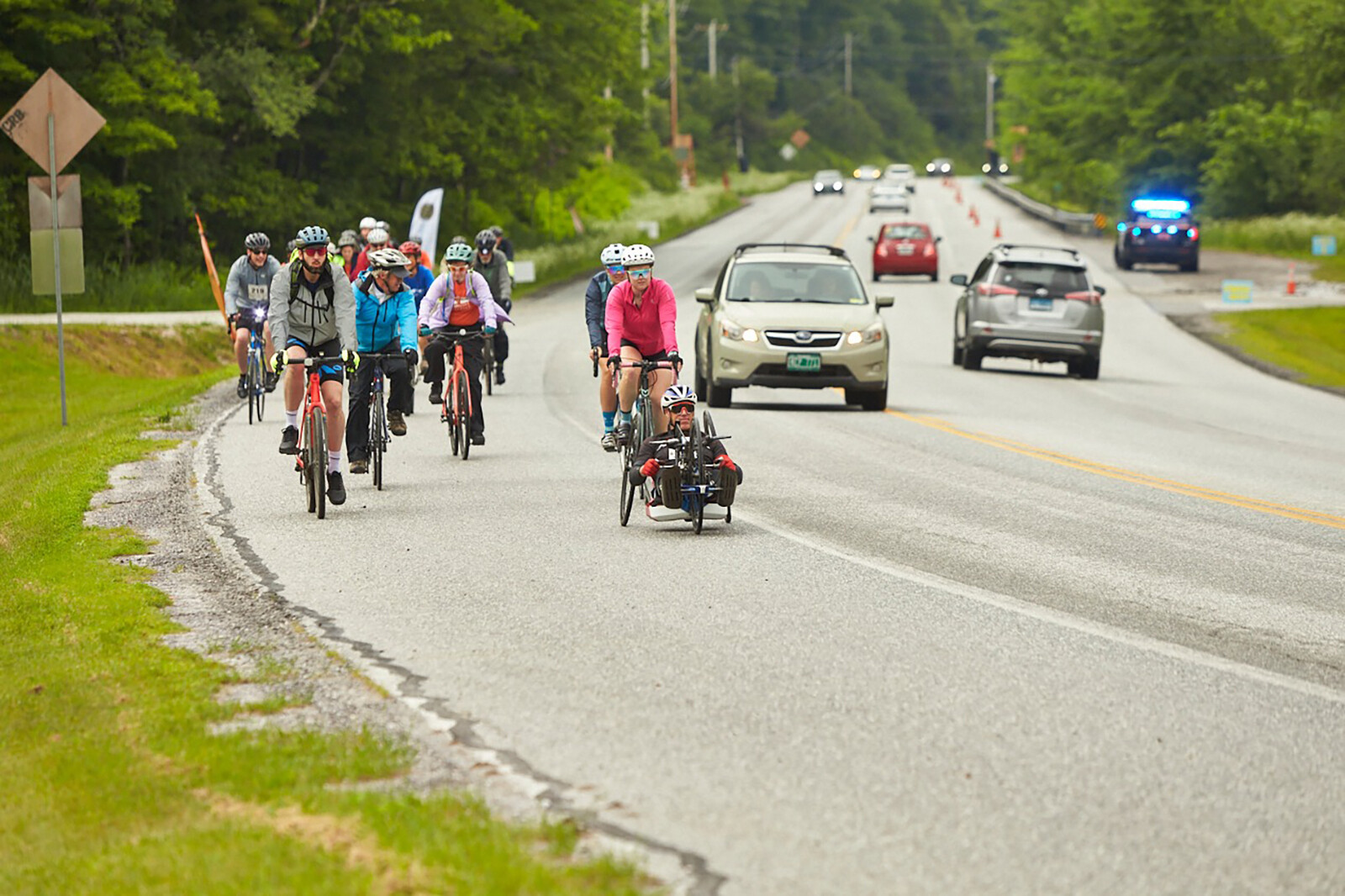 A group of cyclists, including a handcyclist, rides along a road with cars passing by.