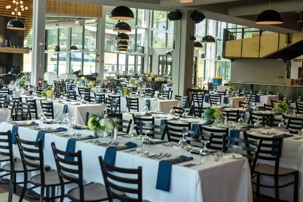 Elegant dining setup with tables adorned in white linens and blue accents, surrounded by windows.