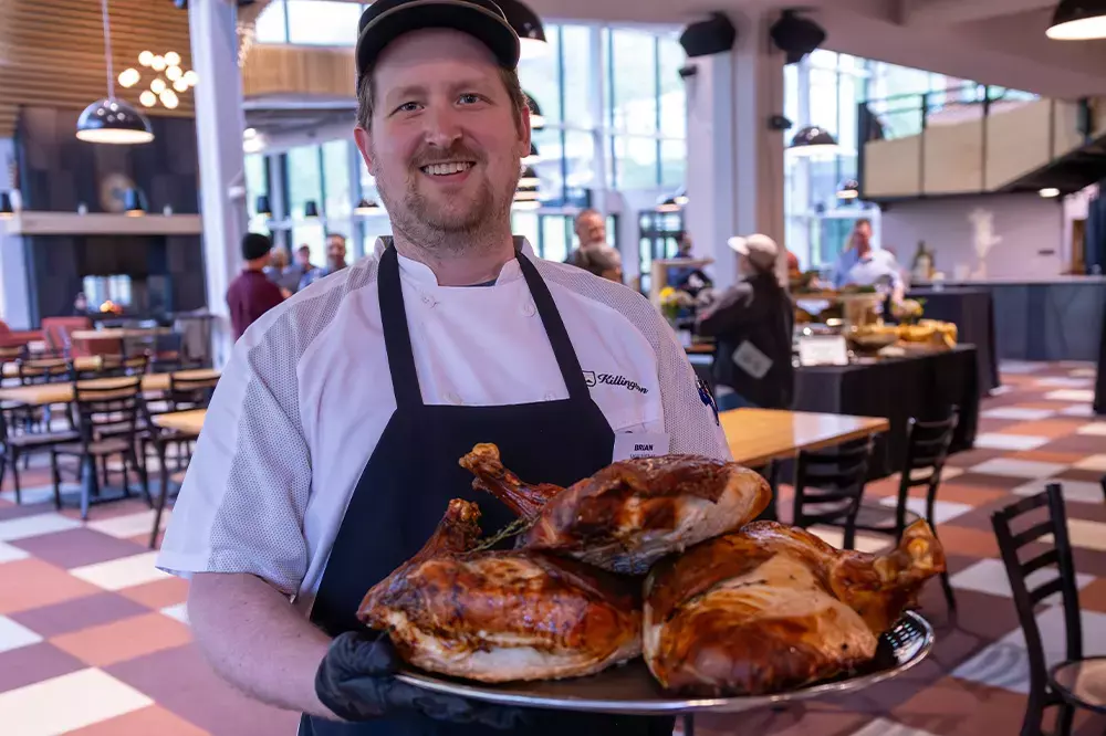 Chef holding a platter of roasted turkeys in a restaurant setting.
