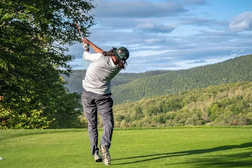 Golfer swings club on a green course with mountains in the background.