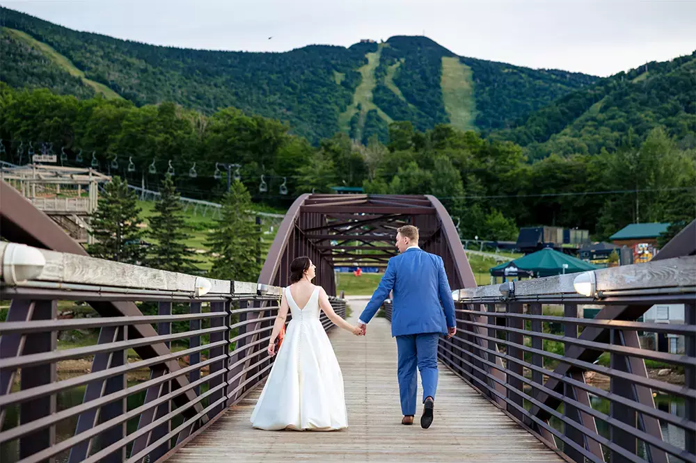 A bride and groom walk hand-in-hand on a bridge with a mountain backdrop at Killington Mountain Resort