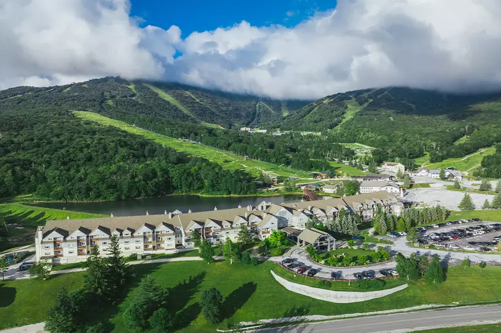 Aerial view of a resort area near mountains, trees, and a lake under a cloudy sky.