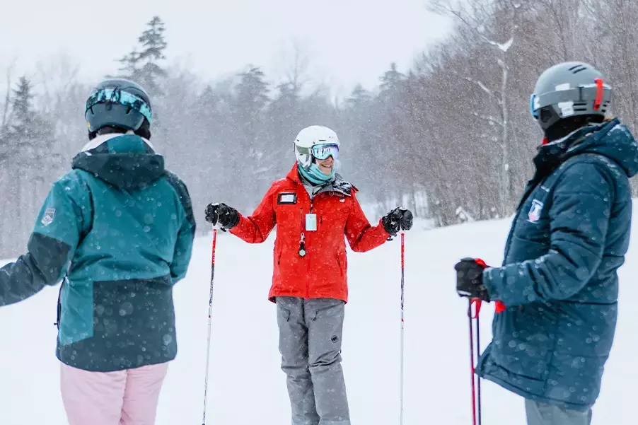 Three skiers in a snowy landscape converse, one wearing a red jacket and helmet.
