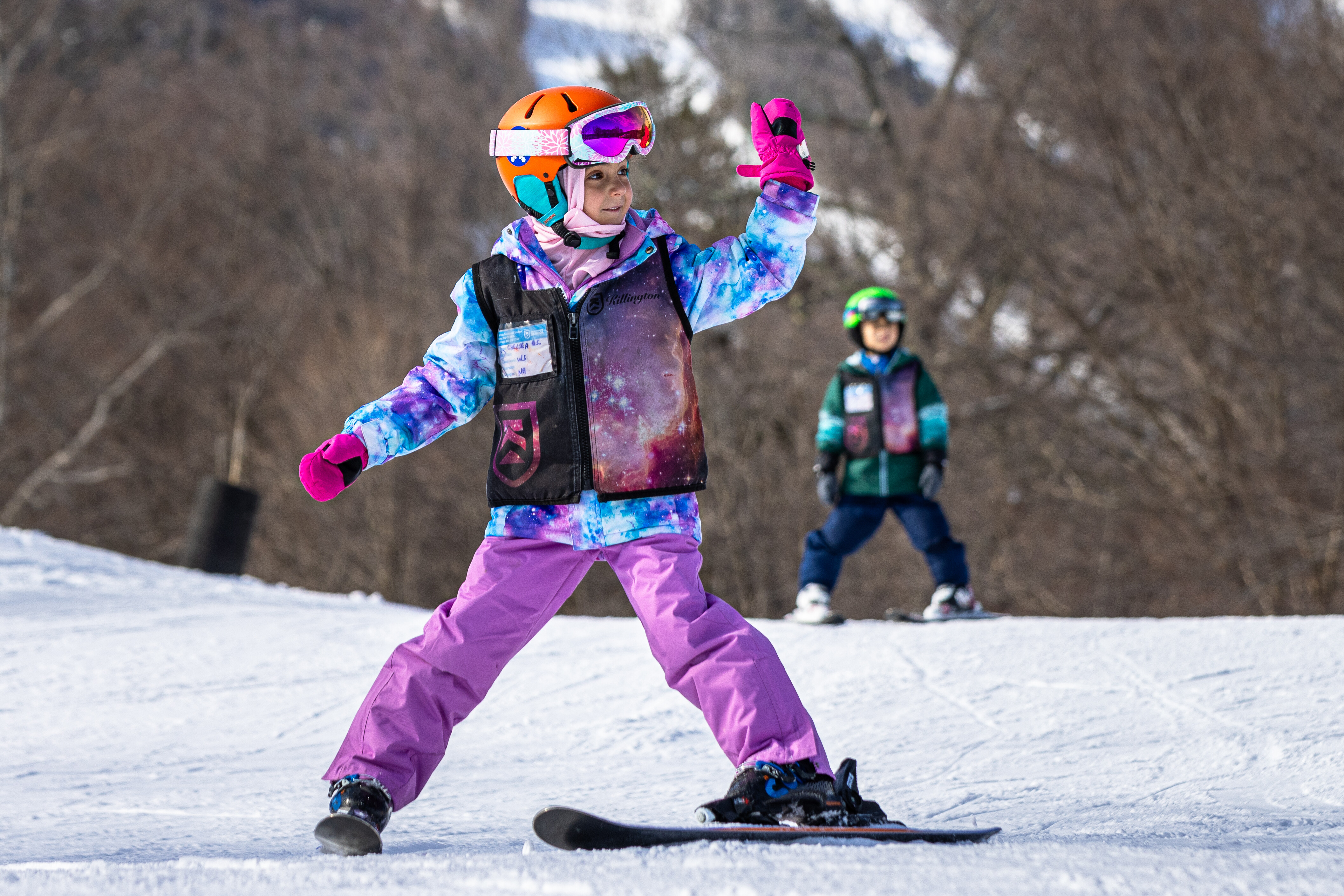 Child snowboarding on a slope, posing with a hand sign, while another child observes.