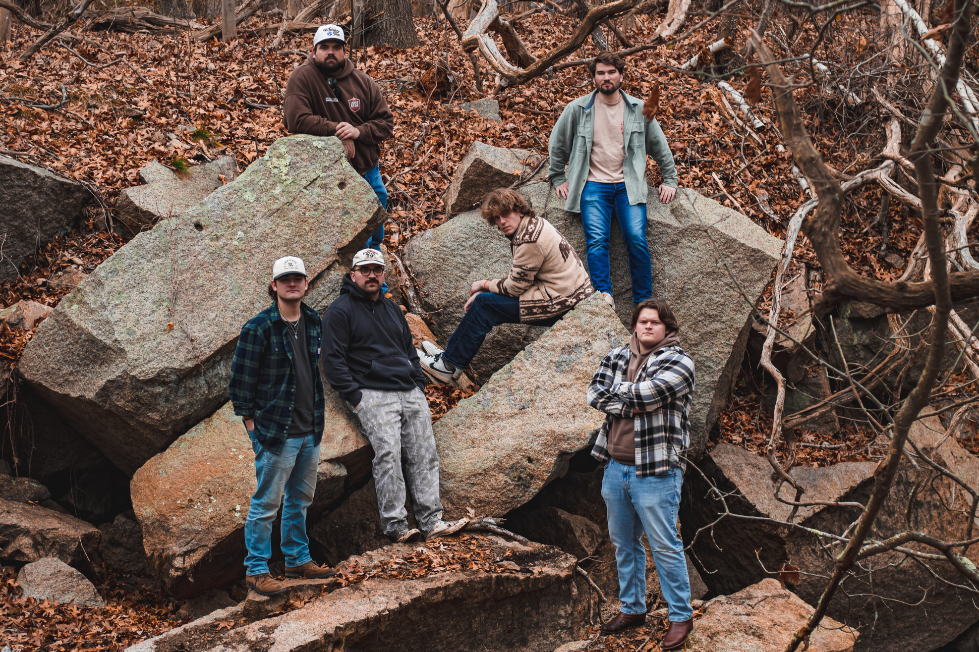 Six men pose on large rocks in a wooded, leaf-strewn area.