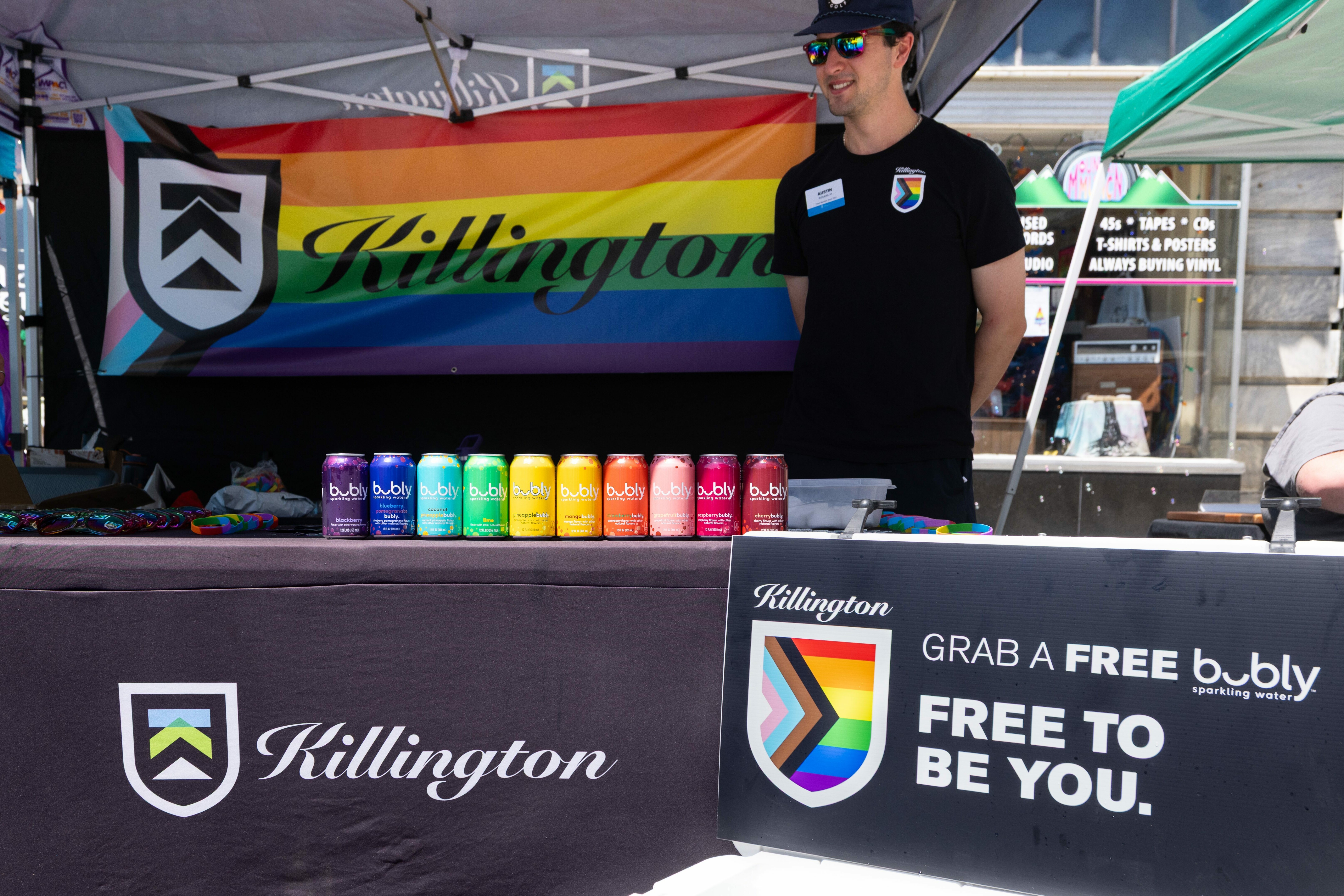 A man stands behind a table with colorful Bubly cans and a Pride-themed banner.