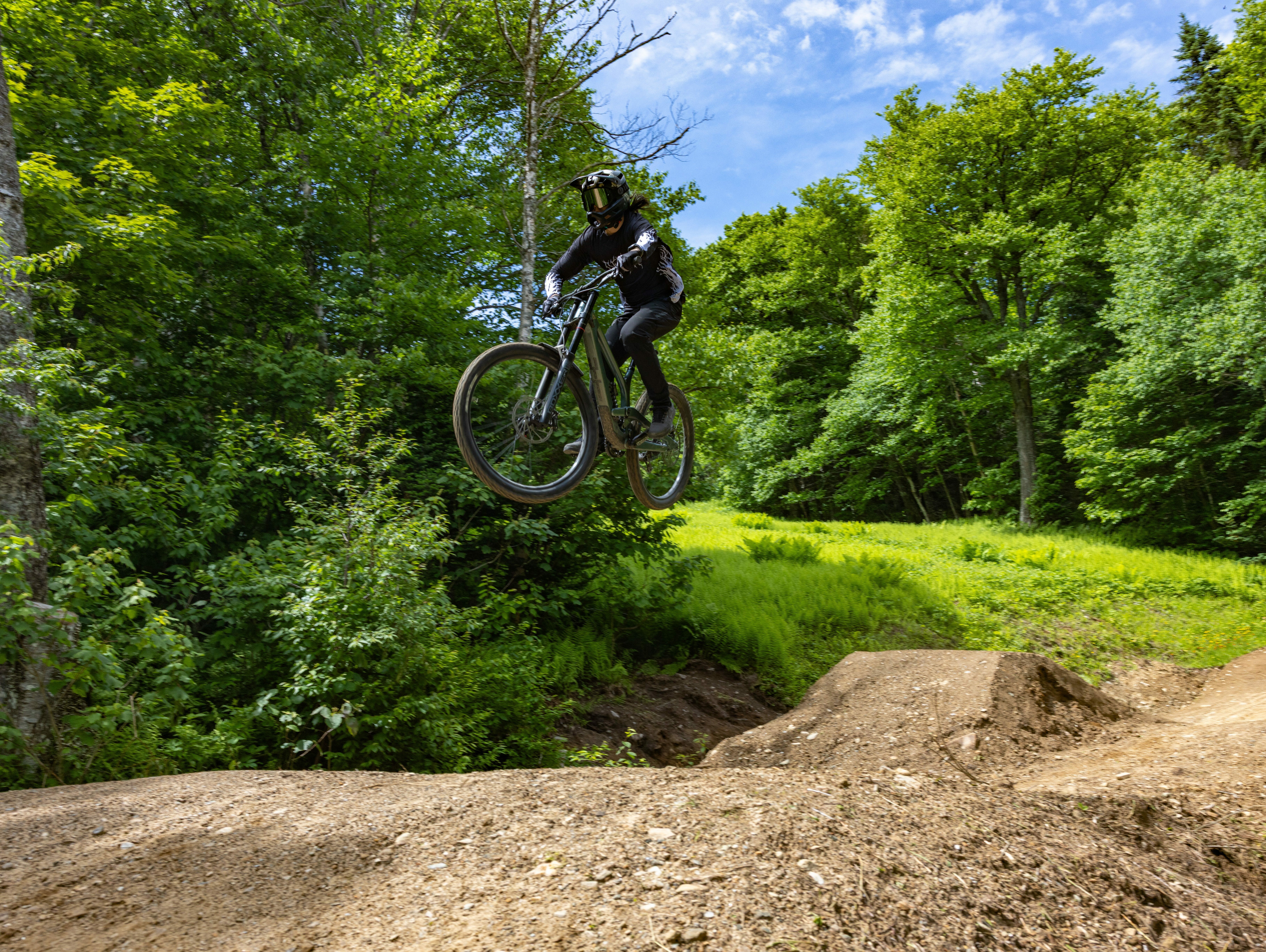 A mountain biker jumps over a dirt ramp in a lush, green forest.