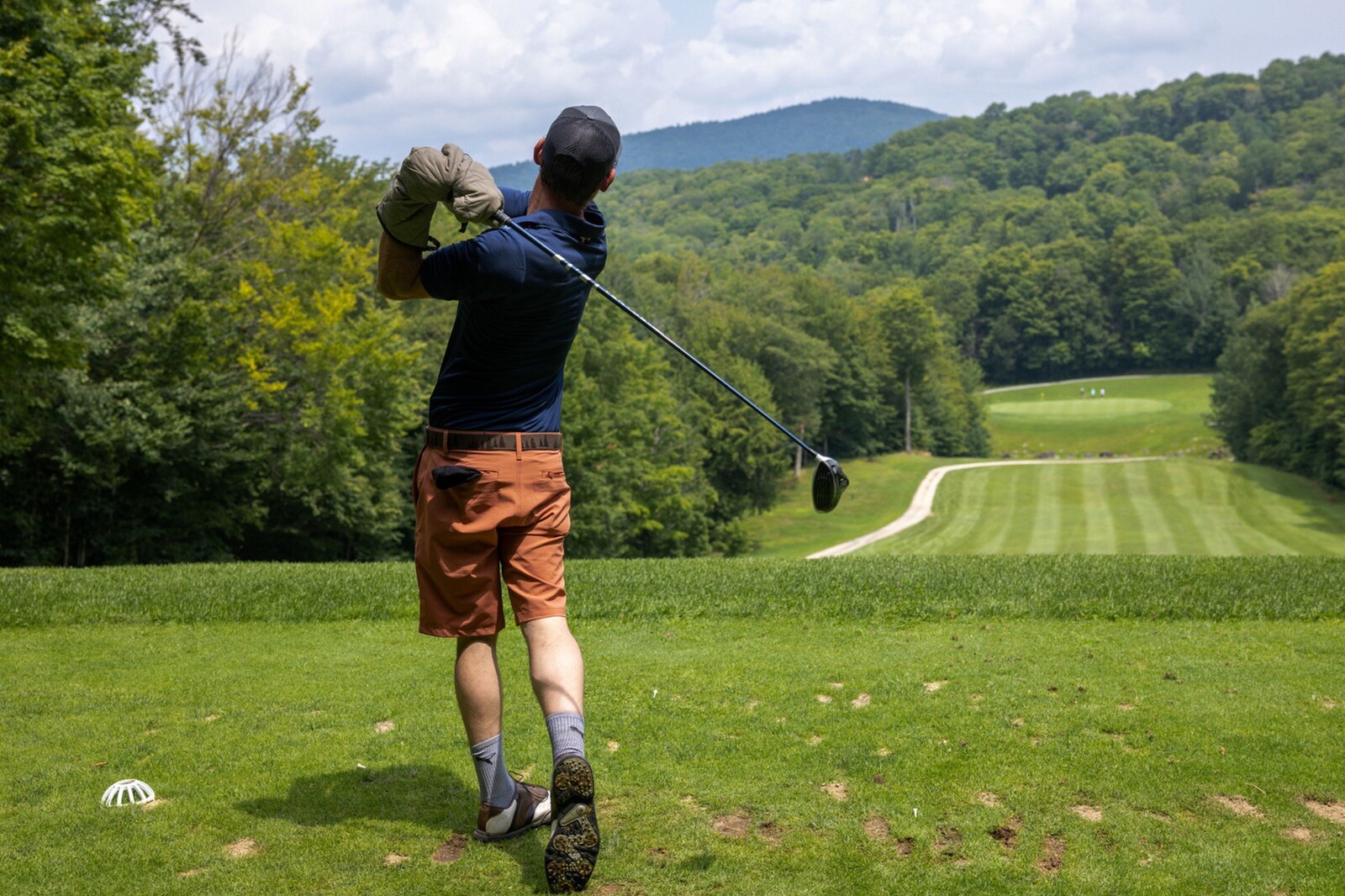A golfer tees off on a lush green course with hills in the background.
