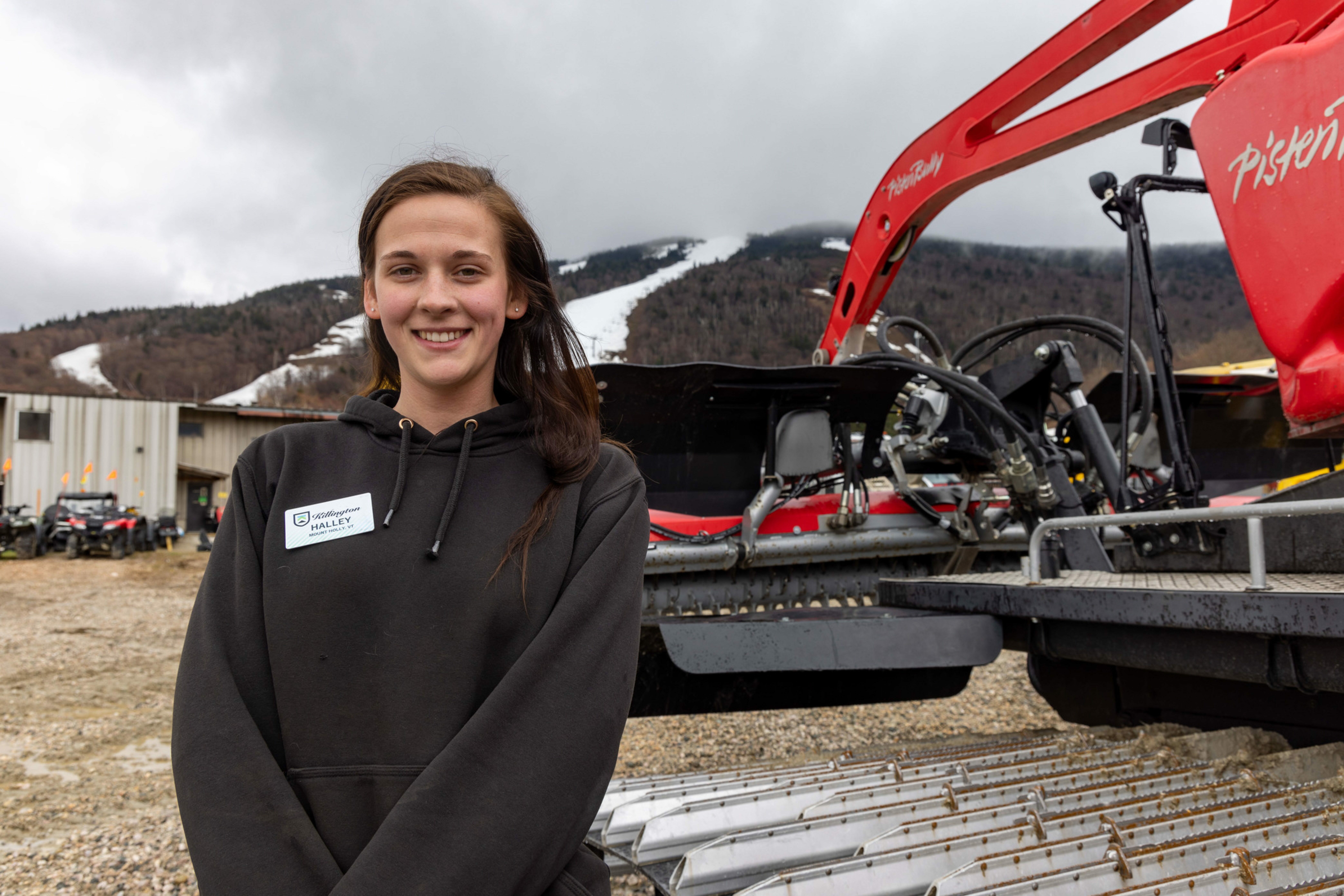 Smiling young woman in a hoodie stands near a red snow grooming machine outdoors.