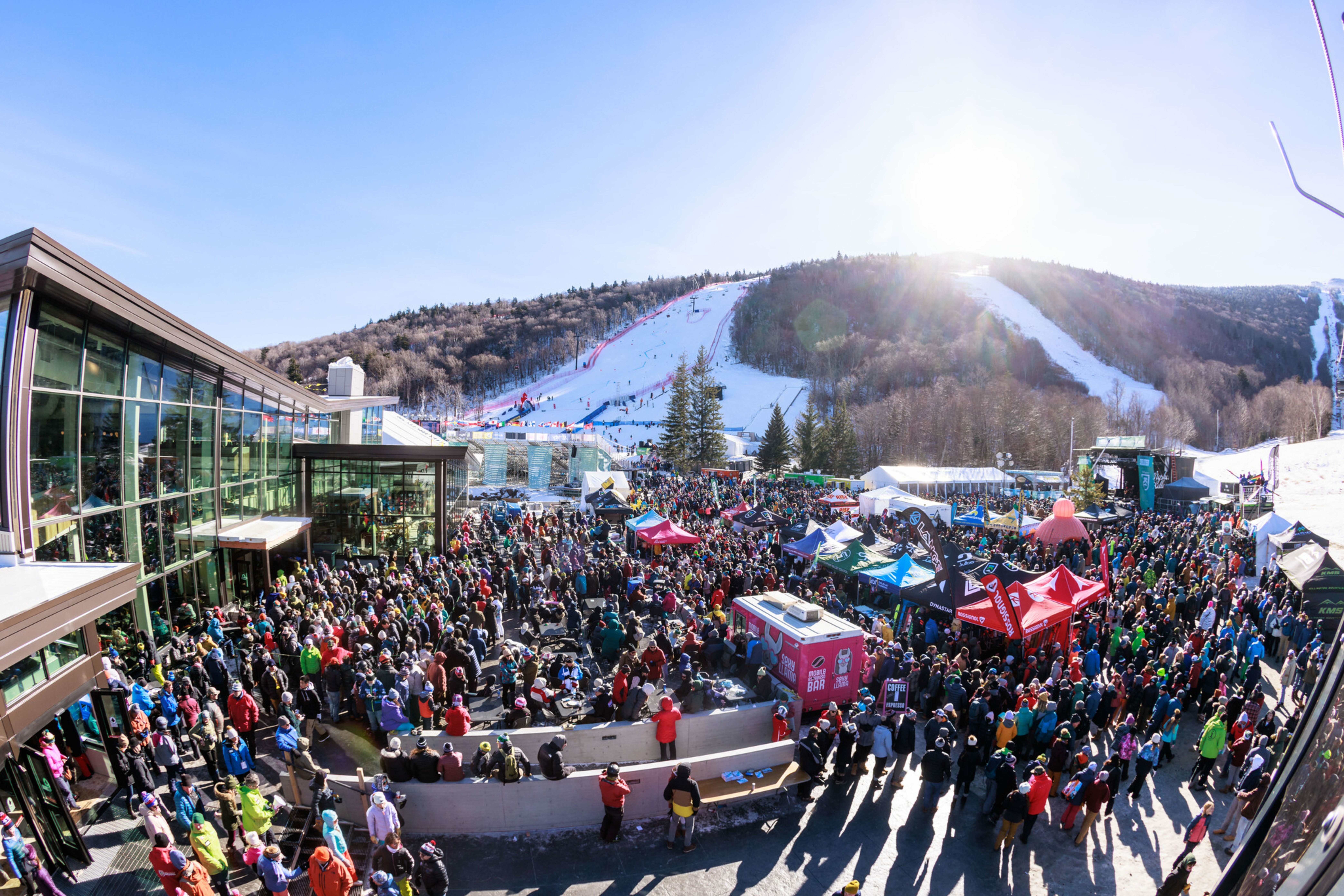 Crowd gathering at a ski resort with mountains and ski slopes in the background.
