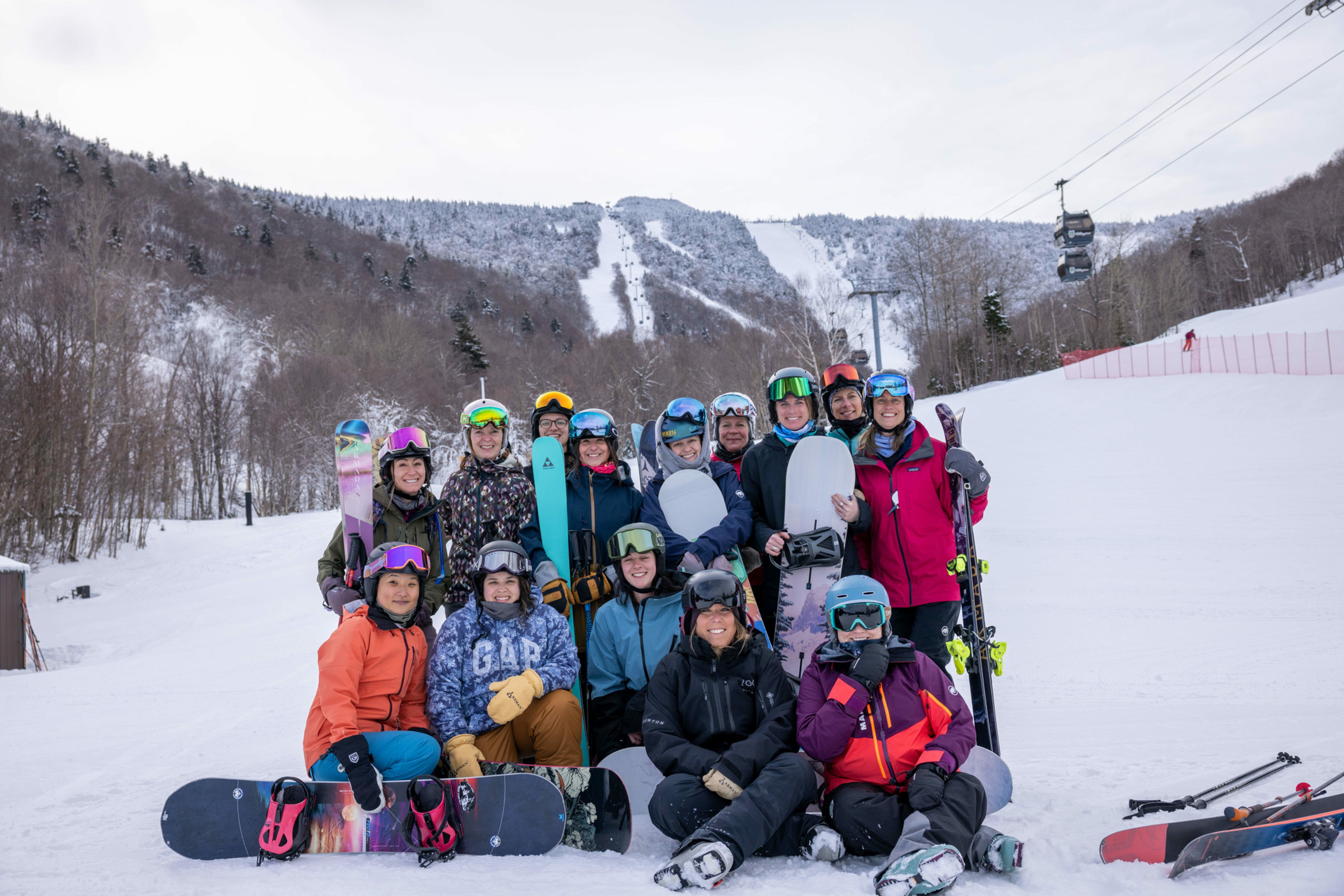 Group of 15 female skiers and snowboarders on a snowy slope, smiling together.