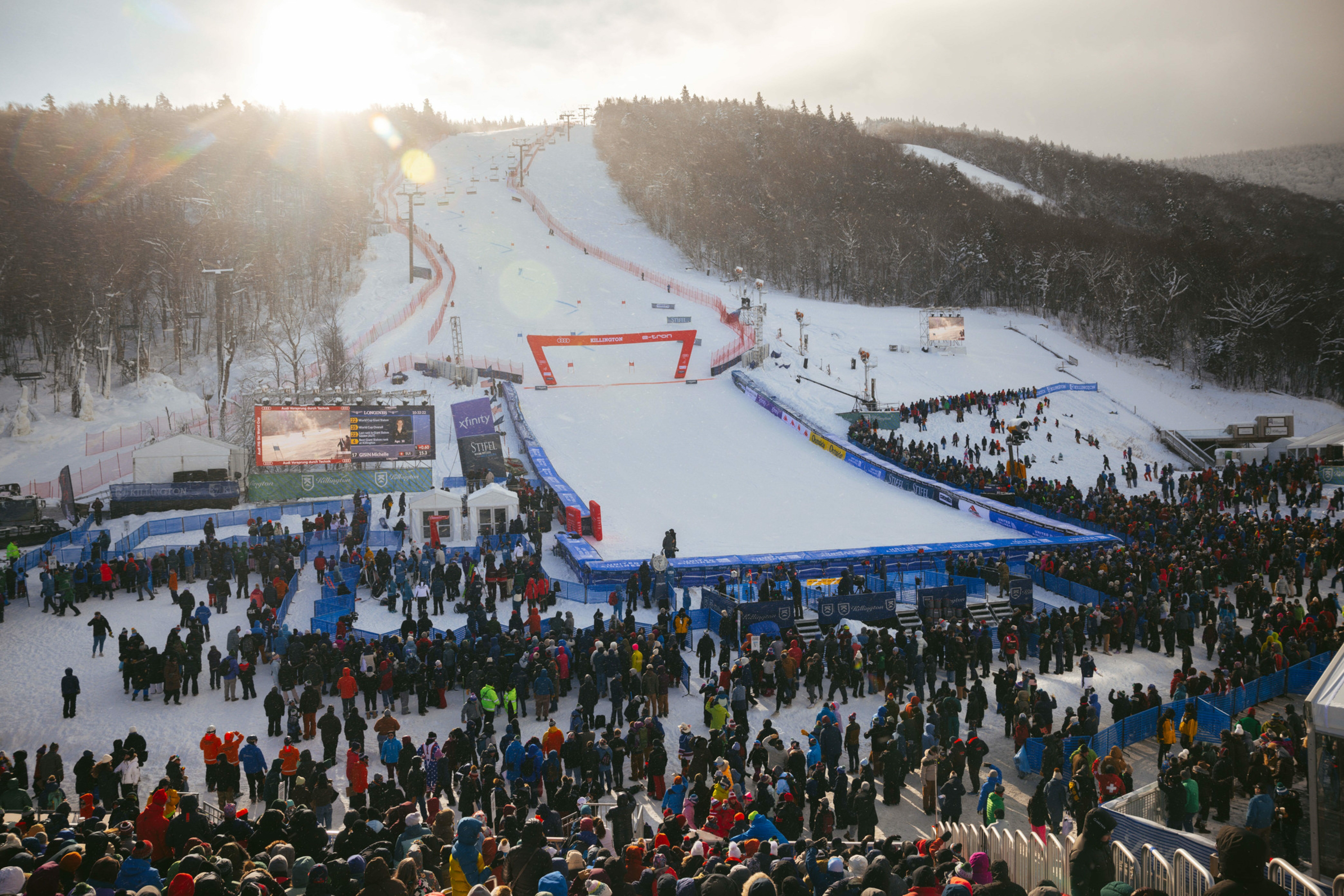 Crowd gathers at a snowy ski slope with an event stage in the foreground.