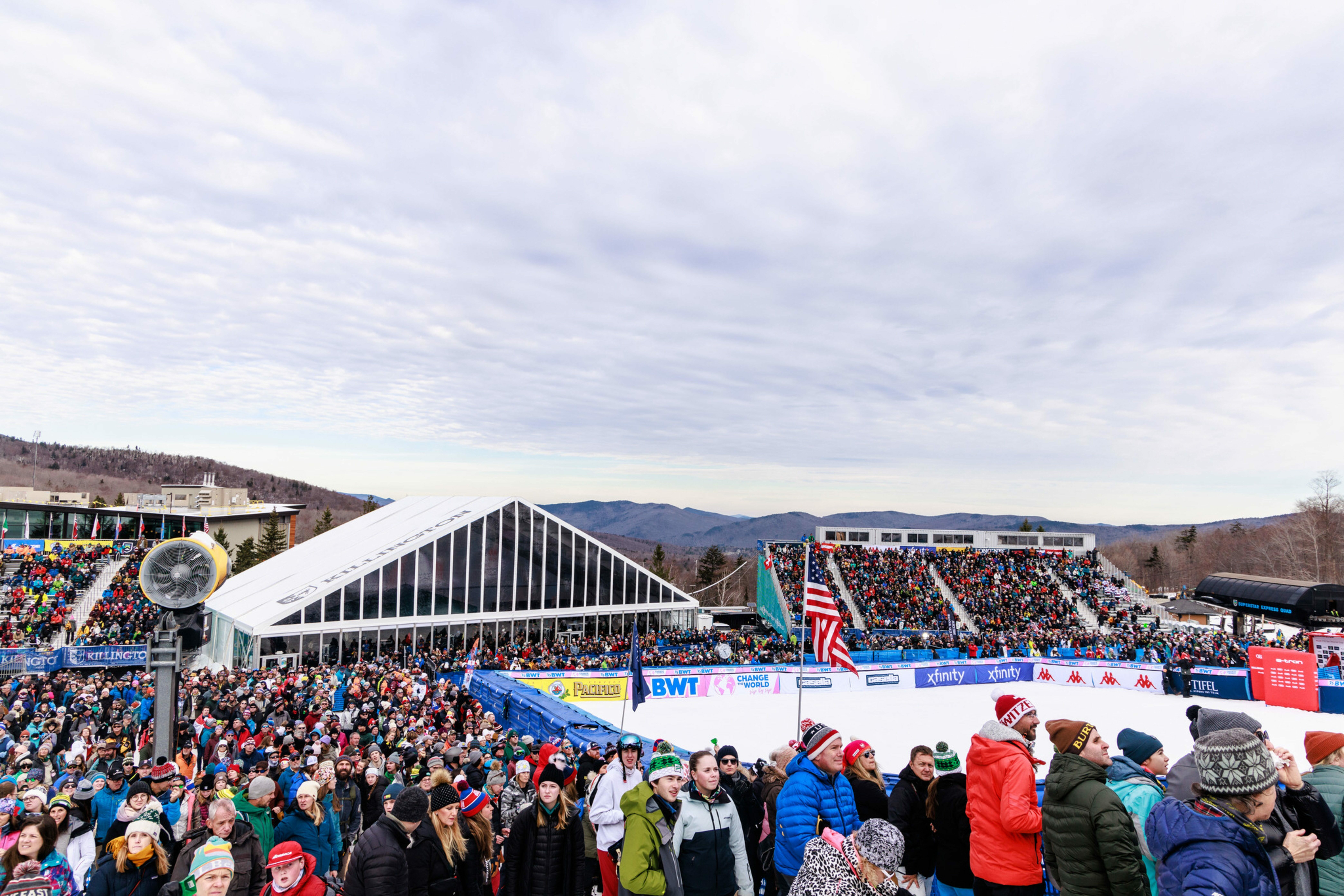 Crowd gathered at a ski event, with mountains and cloudy sky in the background.
