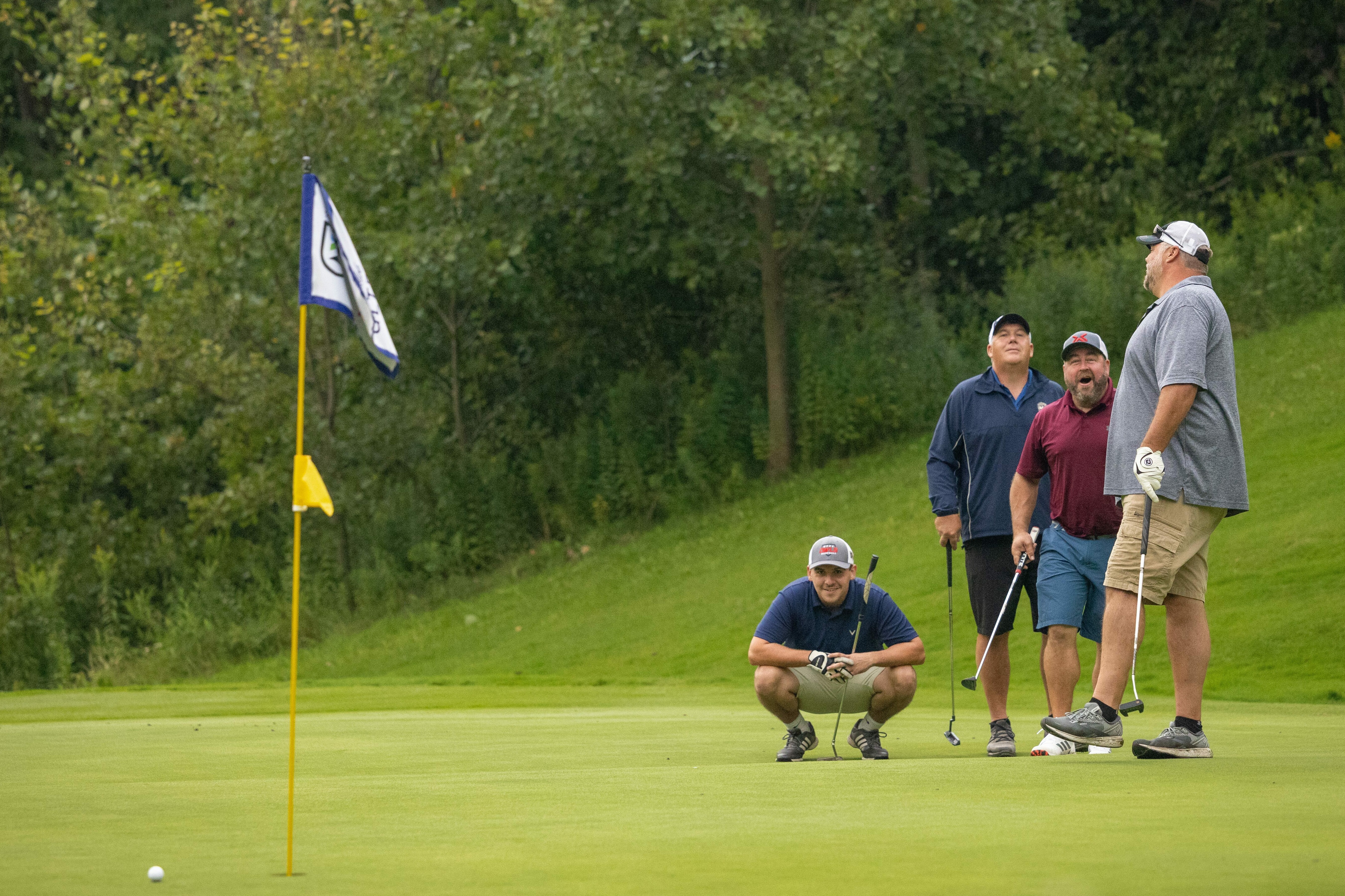 Four men on a golf course observe a putt near the flag. Trees in the background.