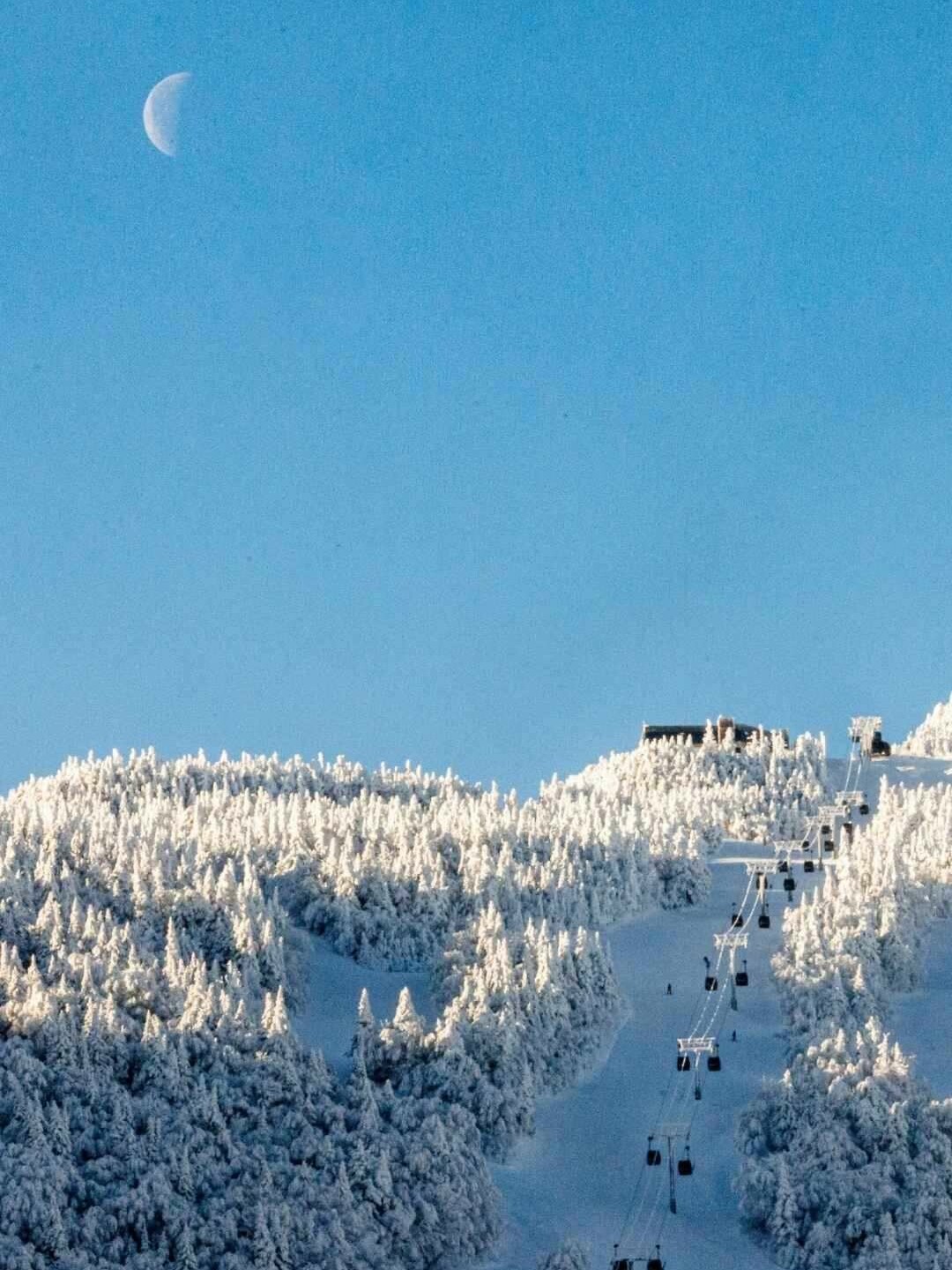 Moon over snow-covered mountains and ski lifts under a clear blue sky.